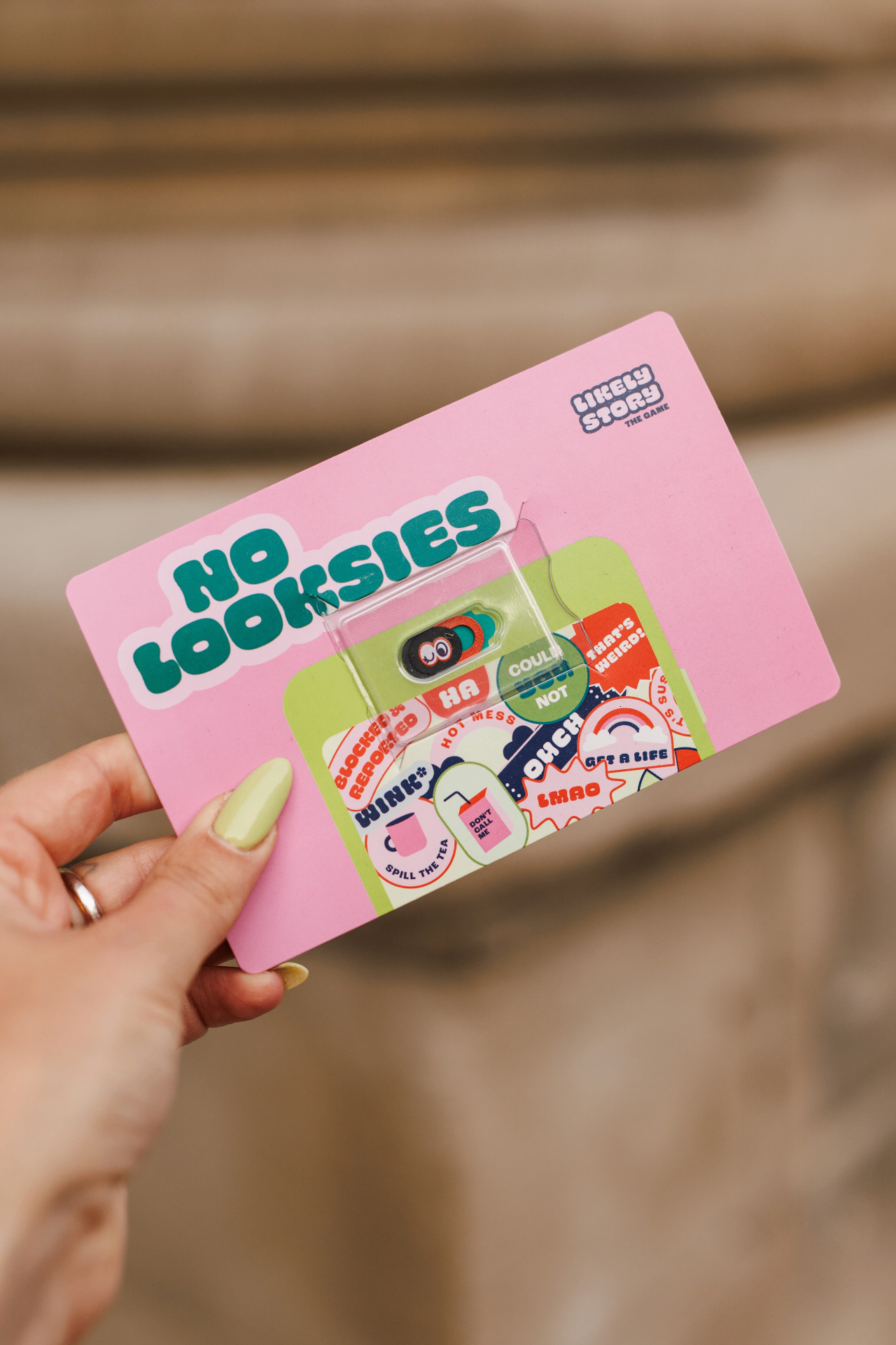 A hand holding a pink 'No Looksies' webcam-cover card from Likely Story, with a sticker sheet of teen-speak reactions like 'wink', 'spill the tea', 'don't call me', and 'get a life'.
