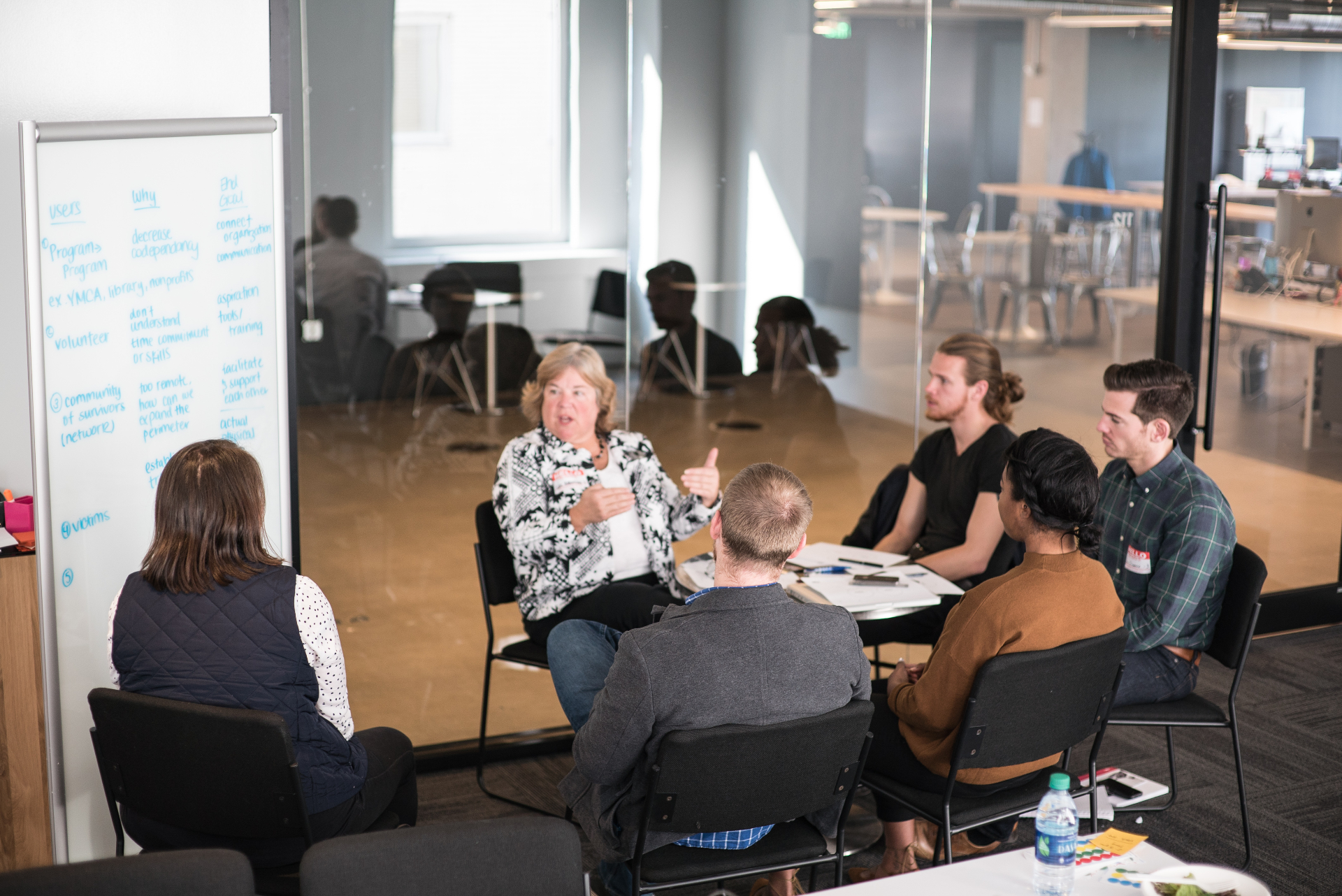 Small-group discussion during the sprint: a subject-matter expert speaking with a team of designers seated around a table, whiteboard notes about users, why, and end goal visible at left.